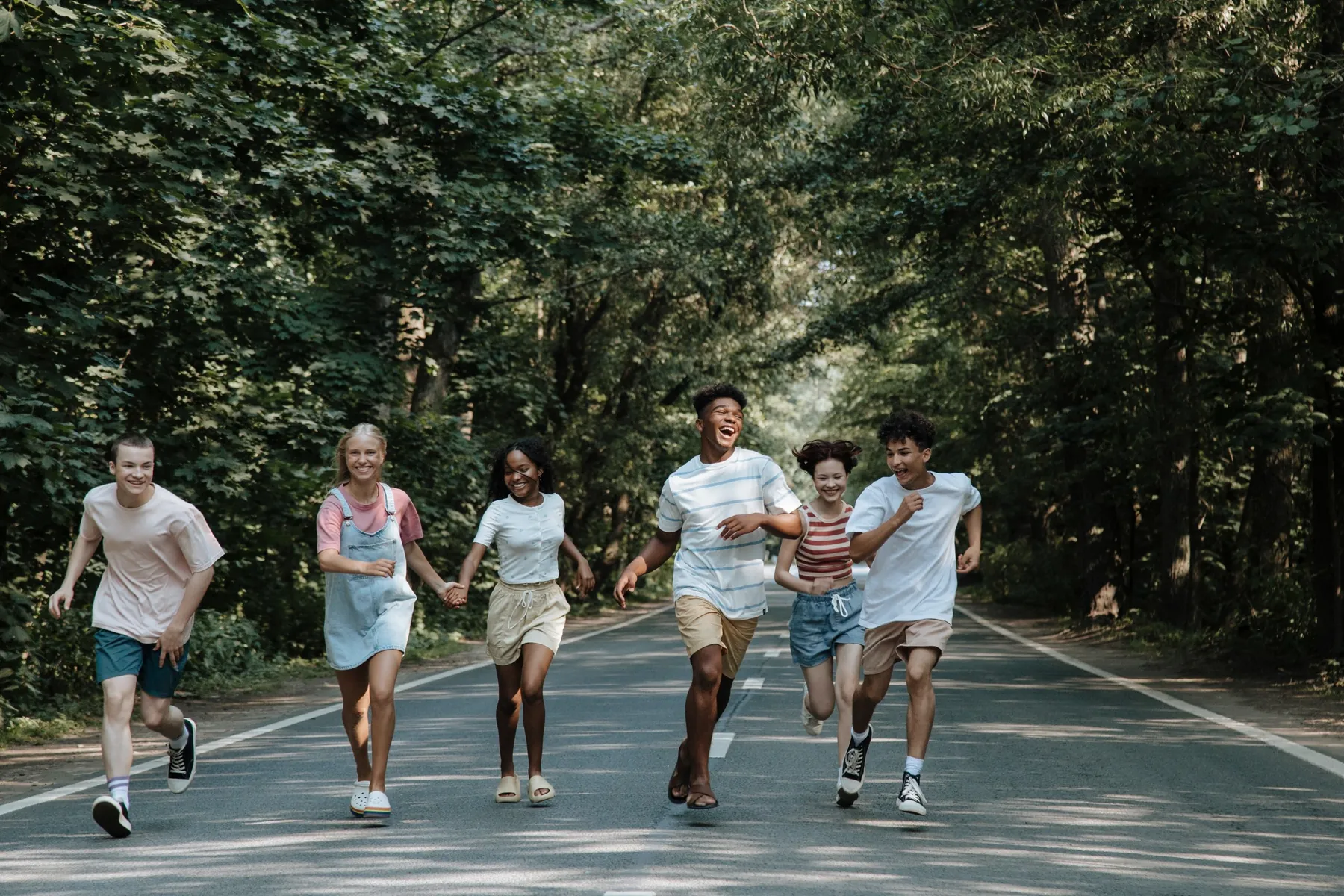A group of teenagers run laughing down a tree-lined road.