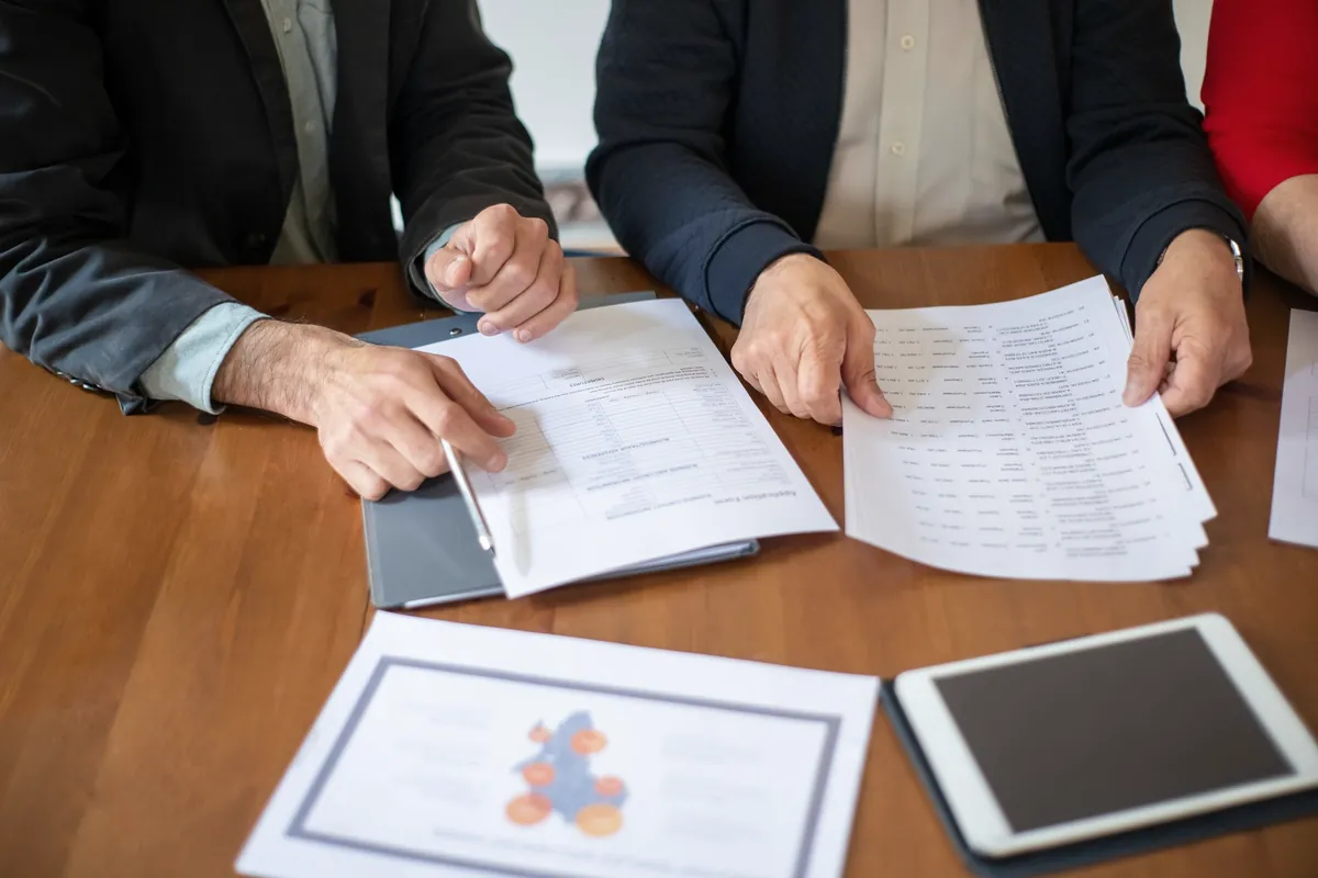 Three people at a wooden table reviewing printed financial documents beside a tablet.