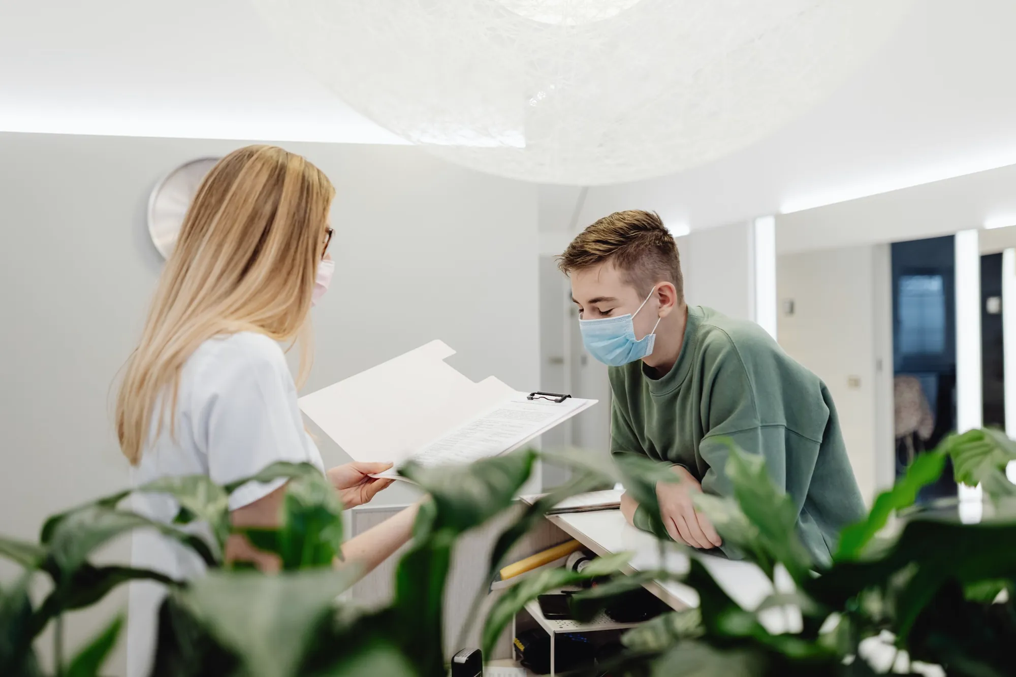 A receptionist at a clinic front desk reviews paperwork with a young patient.