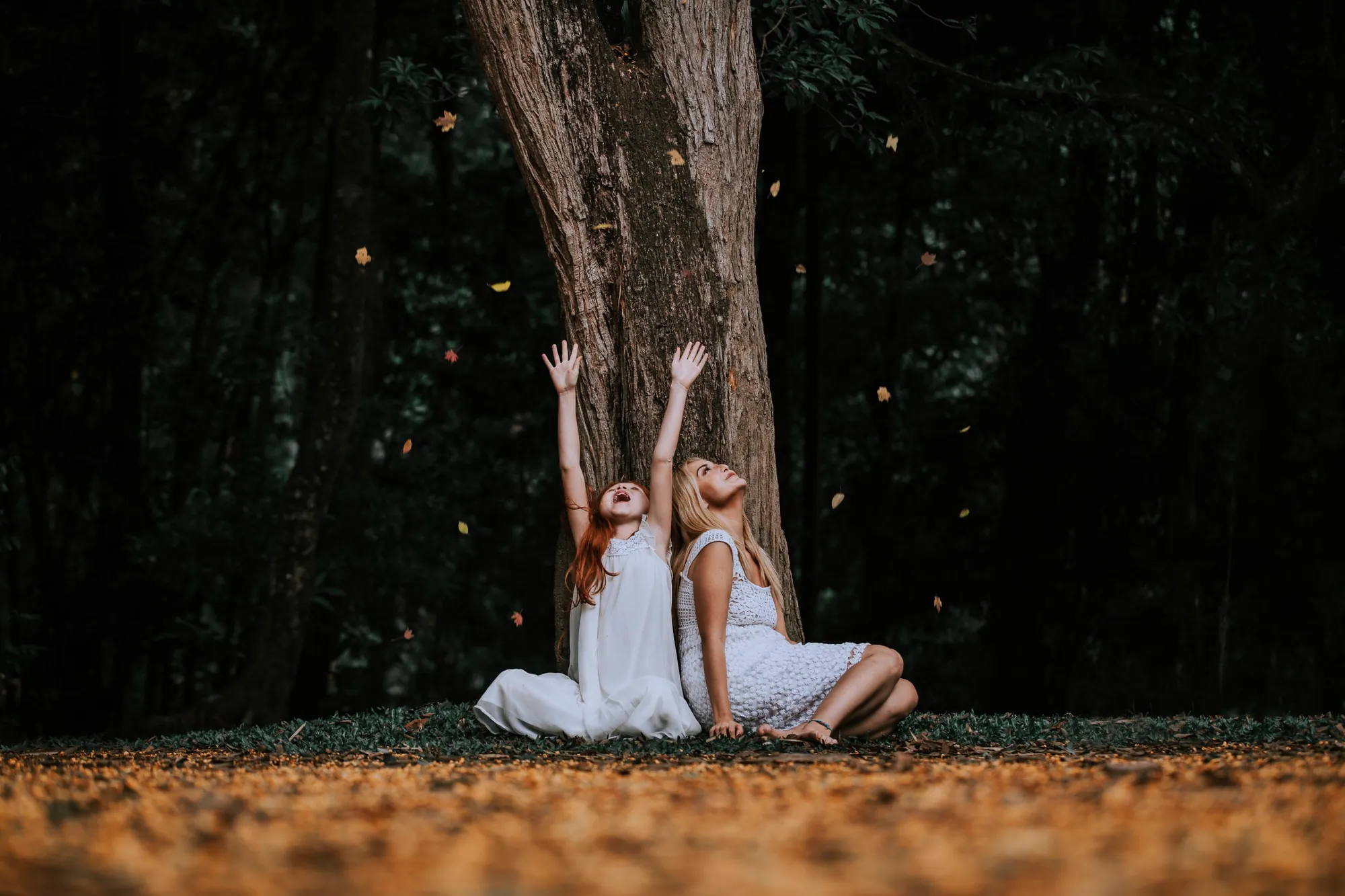 A woman and a girl in white sit against a tree trunk, looking up as leaves fall around them.